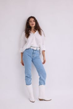 a woman standing in front of a white wall wearing blue jeans and boots with her hands on her hips