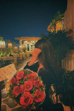 a woman holding a bouquet of red roses in front of a building at night with the lights on