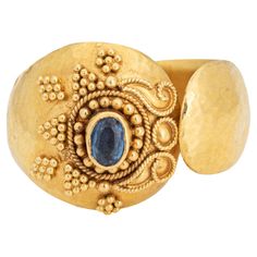 Stylish vintage sapphire ring crafted in 22 karat yellow gold. Small blue sapphire measures 4mm x 3mm. The sapphire is in very good condition and free of cracks or chips. The distinct hand-made ring in bright buttery 22k yellow gold features fine granulation work in a star like pattern and scrolled wire work. Hand hammered details adorn the wide (15.5mm - 0.61 inches) cigar style band. The ring is great worn alone or stacked and makes a nice statement on the hand. The low rise ring (6.5mm - 0.25 Gold Oval Yellow Sapphire Ring, Oval Yellow Sapphire Ring In Gold, Formal Gold Ring With Yellow Sapphire, Gold Sapphire Ring With Gemstone, Formal Gold Sapphire Ring With Yellow Sapphire, Gold Round Yellow Sapphire Ring, Gold Sapphire Rings Hand Forged, Gold Hand Forged Sapphire Ring, Gold Yellow Sapphire Ring, Round