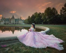 a woman in a pink dress is sitting on the grass near a pond and castle