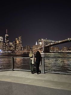 two people standing on a bridge looking at the water and city lights in the background