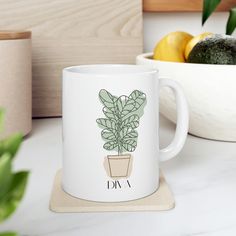 a white coffee mug with a green plant on it next to a bowl of fruit