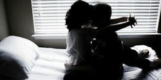 two people sitting on a bed in front of a window with the blinds partially closed