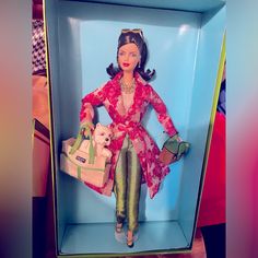 a doll in a box holding a purse and wearing a red dress with green pants