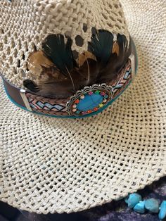 --- This charming straw hat exudes rustic elegance with its natural woven texture and thoughtful embellishments. At the front, a striking turquoise stone serves as the centerpiece, adding a vibrant pop of color. Surrounding the crown is a braided rope band, lending a touch of earthy sophistication. Beautiful brown feathers are artfully arranged, enhancing the hat's organic appeal. Atop the crown, crisscross leather straps provide an intriguing design element, seamlessly blending rugged and refined aesthetics. This hat is perfect for adding a touch of Western flair to any outfit. Bohemian Straw Hat With Short Brim For Kentucky Derby, Bohemian Natural Hat For Kentucky Derby, Bohemian Natural Straw Hat For Kentucky Derby, Western Style Multicolor Sun Hat For Beach, Western Multicolor Sun Hat For Beach, Natural Bohemian Hat For Kentucky Derby, Natural Color Bohemian Hat For Kentucky Derby, Handmade Bohemian Straw Hat For Kentucky Derby, Bohemian Sun Hat For Kentucky Derby And Rodeo