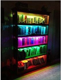 a bookshelf filled with lots of different colored books in a dark room next to a window