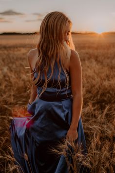 a woman in a blue dress is standing in a wheat field with the sun setting behind her