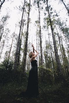 a woman standing in the middle of a forest with her arms raised up to the sky