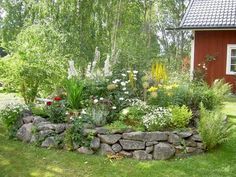 a garden with rocks and flowers in the grass next to a red house on a hill