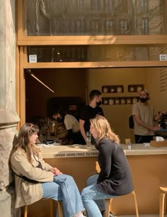 two women sitting at a counter in front of a coffee shop with people standing around