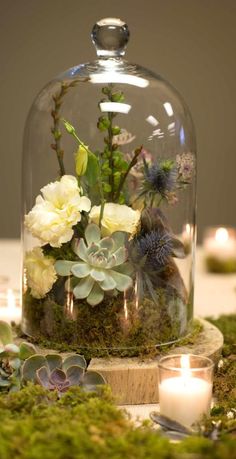 a glass clochel with flowers and greenery under it on a table next to candles