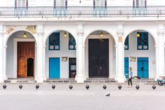 a person walking in front of a white building with blue doors and black birds on the sidewalk