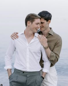 two men standing next to each other in front of the ocean with their arms around each other