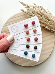 a person is holding four different colored buttons in front of a dried plant on a wooden plate