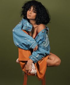 a woman in an orange dress and jean jacket sitting on a stool