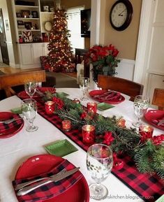 the table is set for christmas dinner with red and green placemats, plaid napkins, silverware, candlesticks and greenery