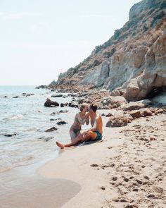 two people sitting on the beach next to each other near the water and rocks in the background