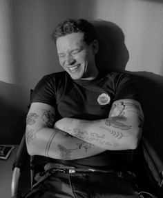 a man sitting in a chair with his arms crossed and tattoos on his arm, smiling