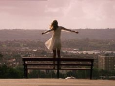 a woman standing on top of a bench with her arms spread wide open in front of the city