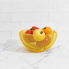 a yellow mesh bowl filled with tomatoes and oranges on a marble countertop in front of a white brick wall