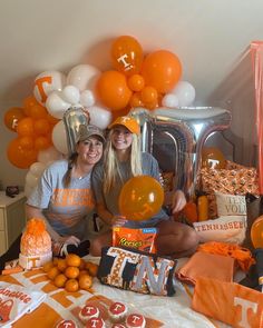 two women sitting on a bed with orange and white balloons in the air behind them