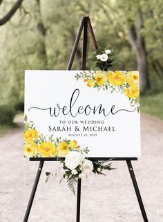 a welcome sign with yellow flowers and greenery on it is standing in the middle of a dirt road