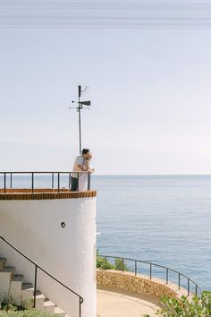 a man and woman standing on top of a white building next to the ocean with their arms around each other