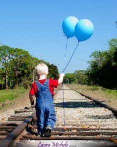 a young boy holding two blue balloons while walking on train tracks