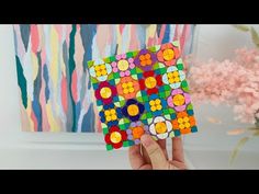 a person holding up a piece of art made out of colored paper with flowers in the background