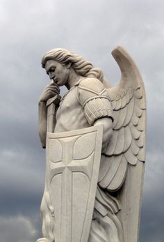 a statue of an angel holding a shield and talking on a cell phone in front of a cloudy sky