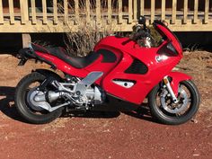 a red motorcycle parked on top of a dirt field next to a wooden fence and shrubbery