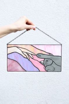 a hand holding a stained glass hanging from a chain on a white wall with a pink and blue background