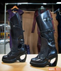 a pair of black boots sitting on top of a wooden table in front of clothes