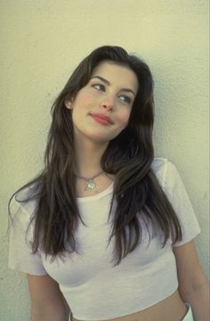 a woman with long dark hair wearing a white shirt and posing for the camera in front of a wall