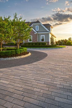 a brick driveway with trees and bushes in front of a large house at sunset or dawn