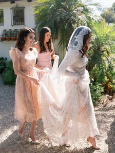 three women in dresses and veils are standing outside together, one is holding the bride's dress