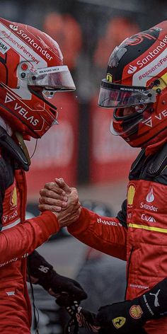 two men in red racing suits shaking hands on the track with helmets on their heads