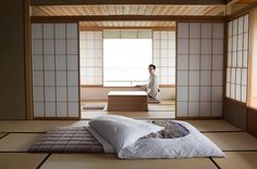 a man sitting on top of a bed in a room next to a table and window