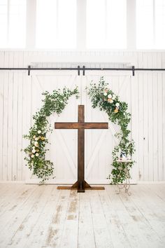 a cross decorated with flowers and greenery in front of a white wall