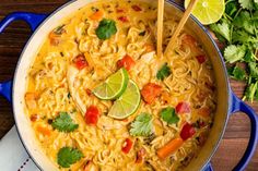 a blue pot filled with noodles and vegetables on top of a wooden table next to cilantro