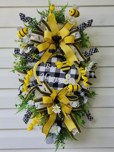 a yellow and black wreath hanging on the side of a white house with bees, daisies and flowers