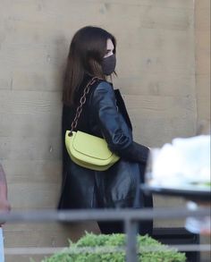 a woman wearing a face mask and carrying a yellow handbag while standing next to a wall