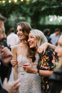 two women hugging each other while standing next to others at an outdoor party with string lights in the background