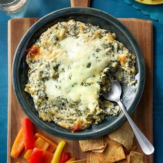 a bowl filled with spinach and cheese on top of a cutting board next to chips