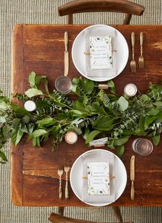 a wooden table topped with white plates and greenery next to forks and knifes