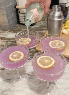 a person pours a drink into three glasses filled with purple liquid and lemon slices