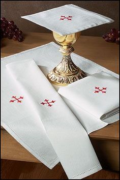 two white napkins with red crosses on them sitting next to a gold cup and saucer