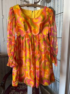 This fabulous dress by Joyce Palmer is made of medium weight golden yellow fabric with a sheer polyester print overlay in a large paisley type pattern, in shades of orange, yellow, green and light and dark pink. The sleeves are sheer with gathers at the cuff (please note that the elastic is no longer stretchy). The dress has darts at the bust and an empire waist, and fastens with a hook and eye and long zipper in the back. Marked size 7/8. The measurements, taken with the dress lying flat, are: Summer Party Dress With Vintage Print, 1970s Style Orange Summer Dress, Retro Orange Dresses With Vibrant Print, Retro Orange Dress With Vibrant Print, 1970s Style Mini Summer Dress, Vintage Orange Mini Dress For Spring, Green Print Dress, Wardrobe Shopping List, 1960s Dresses