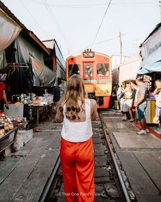 a woman standing on train tracks in front of an orange and yellow train at a market