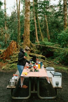 a woman standing at a picnic table with food on it in the middle of a forest